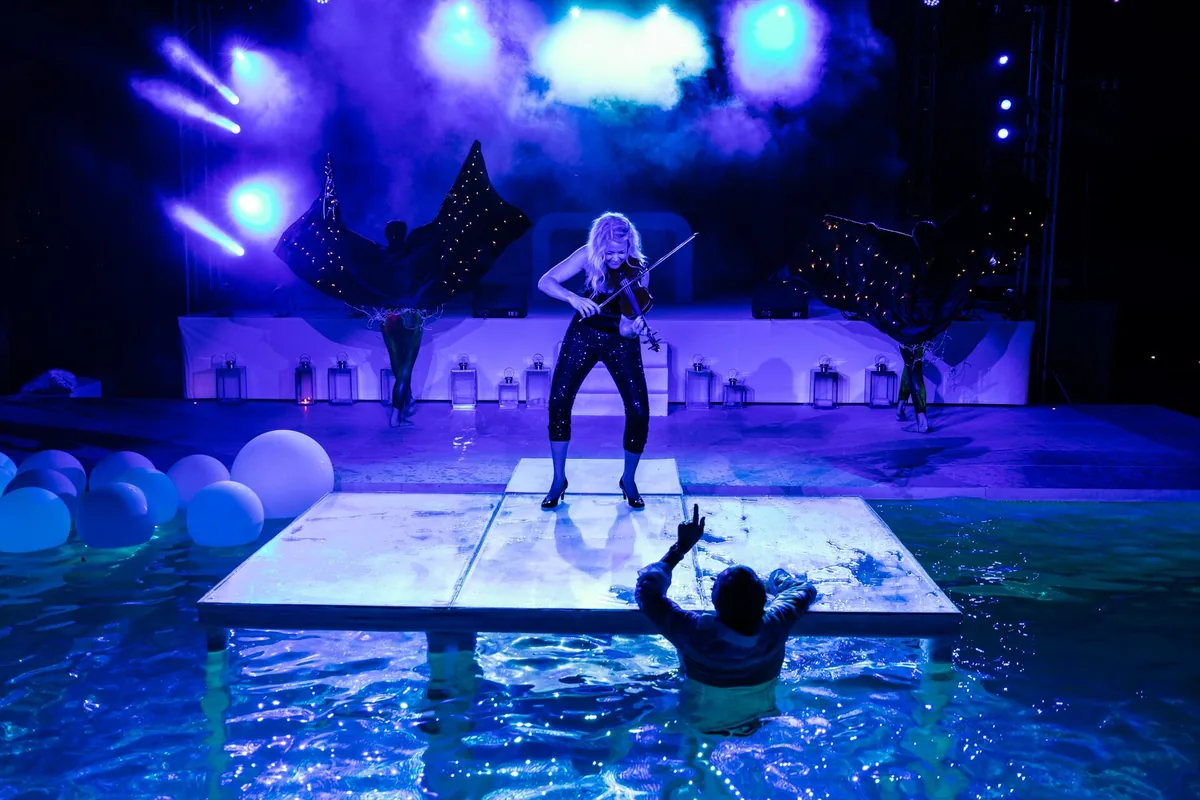 Aja Humm playing the violin on a lit platform in a swimming pool, with dancers and blue stage lights behind her